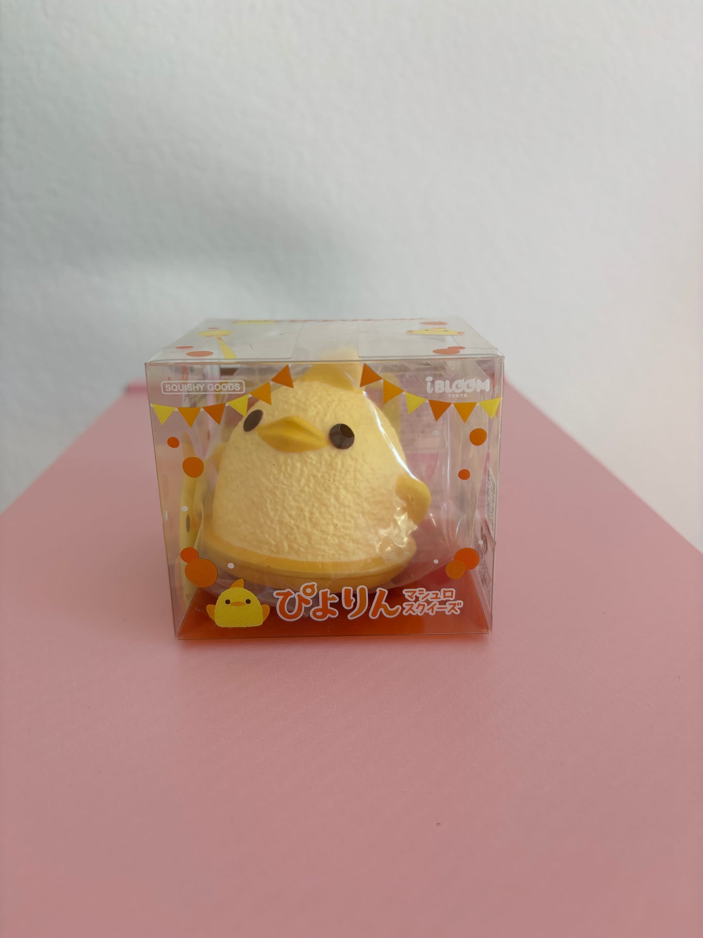 A small yellow foam squishy shaped like a duck, packaged in a clear plastic box with orange and white graphics, set against a white and pink background.