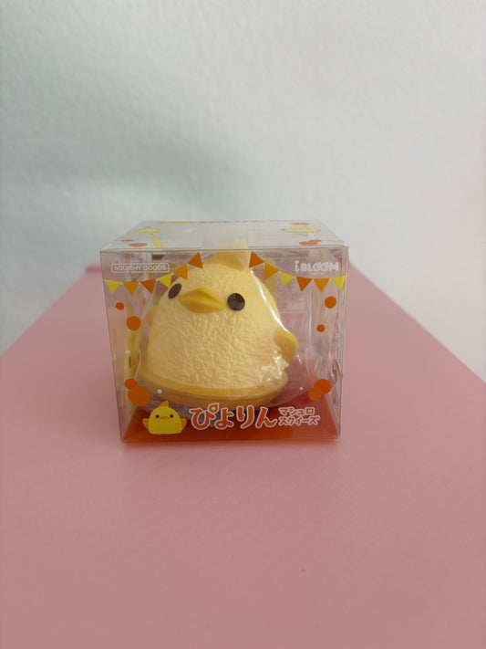 A small yellow foam squishy shaped like a duck, packaged in a clear plastic box with orange and white graphics, set against a white and pink background.