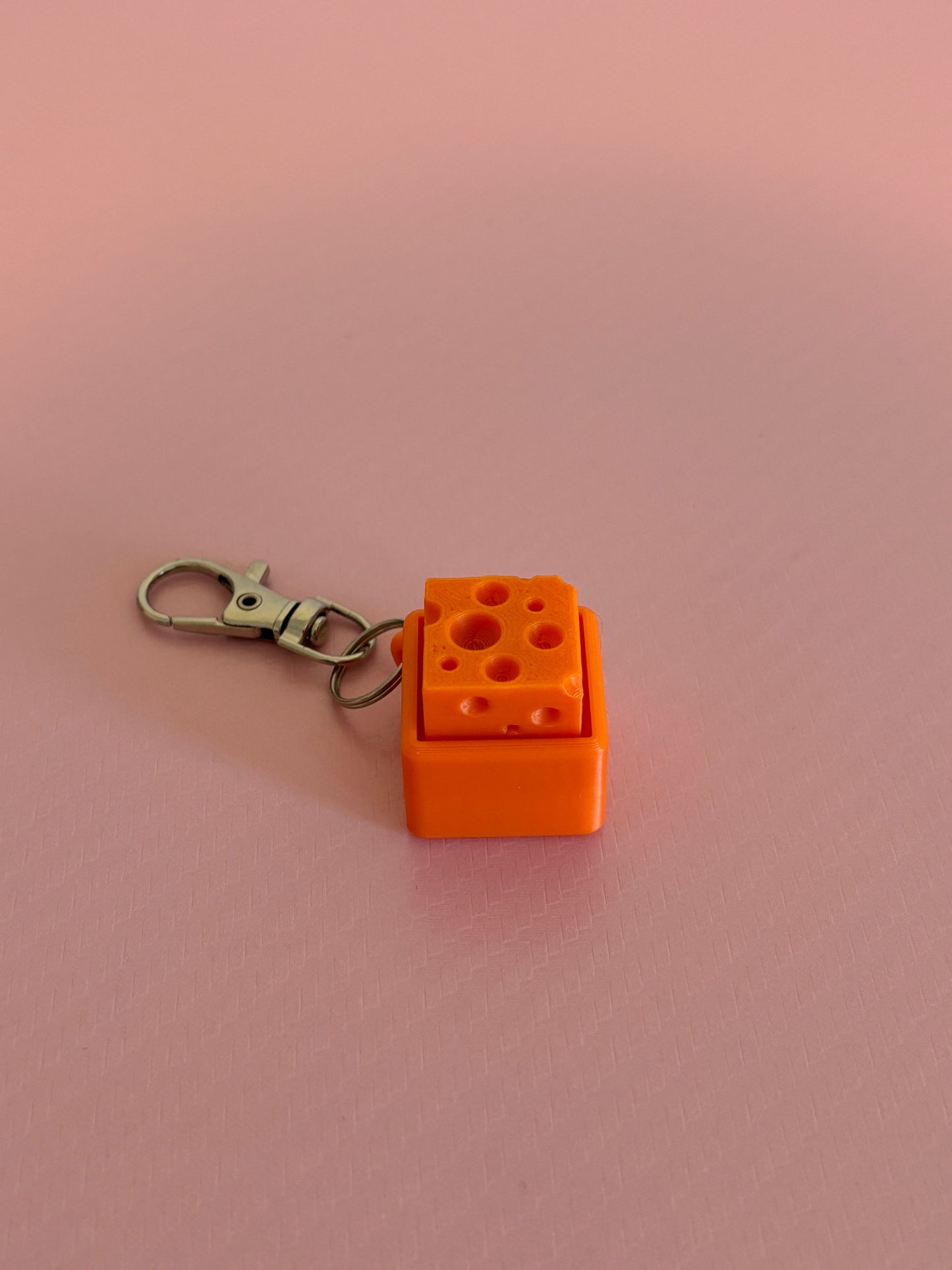 Miniature cheese-shaped keychain clicker in orange attached to a silver metal lobster clasp, displayed on a pink background.