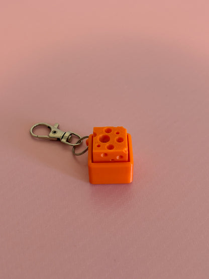 Miniature cheese-shaped keychain clicker in orange attached to a silver metal lobster clasp, displayed on a pink background.
