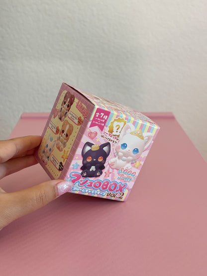 A hand holding a squishy blind box featuring cute cartoon animal illustrations, including a black cat and a white cat on a pink tabletop against a white wall.