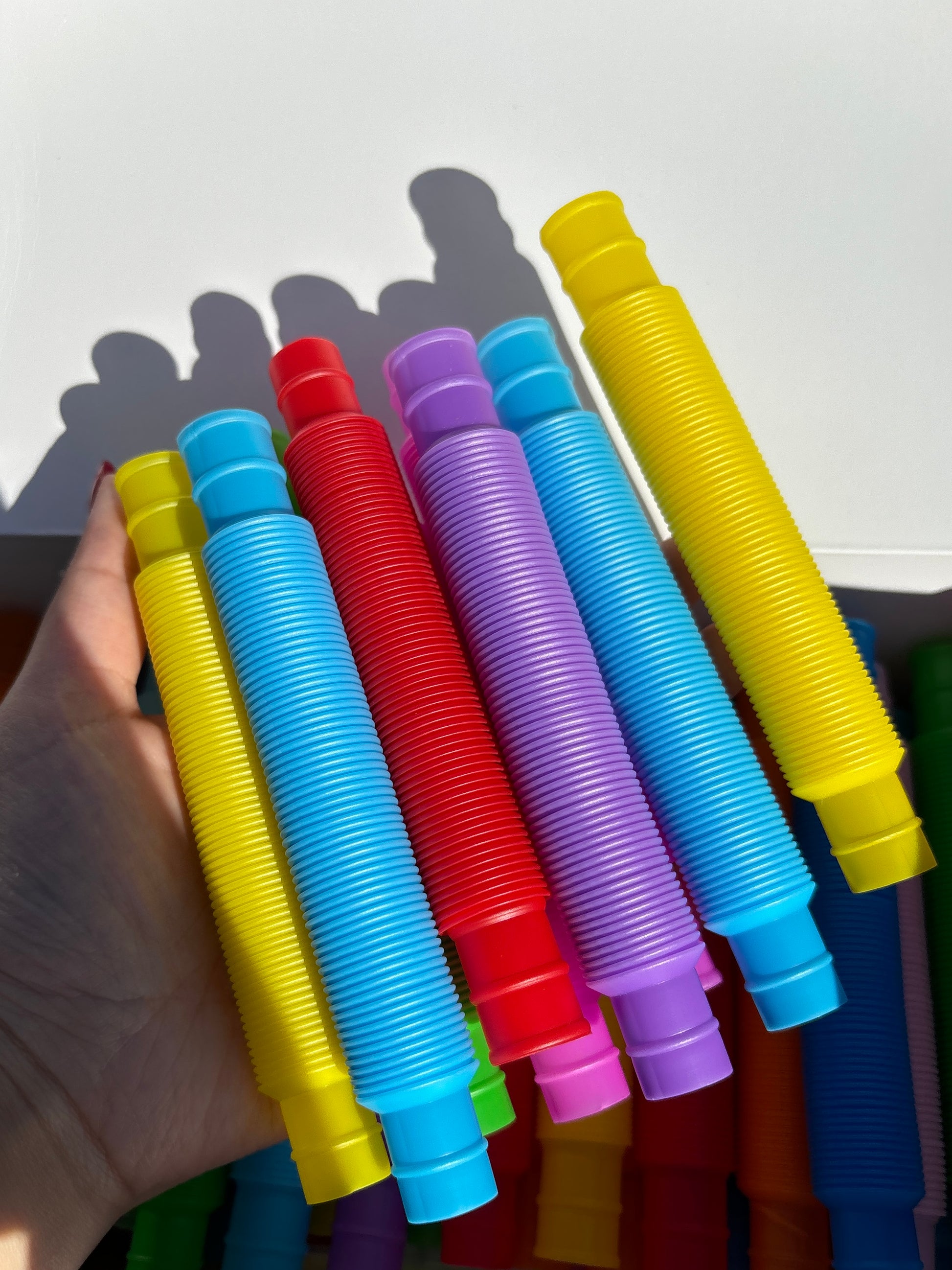 Variety of pop tube color ways including yellow, sky blue, red, purple, and pink being held up in front of other pop tubes