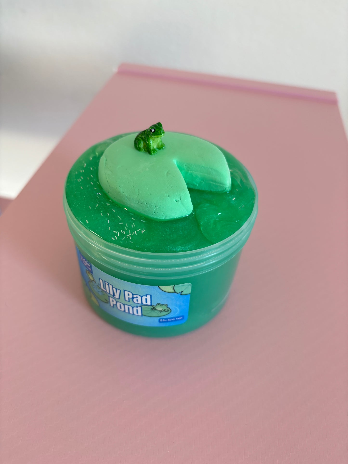 Green Jelly slime in container labeled “Lily Pad Pond” with clay lily pad and frog charm on top, in front of pink background