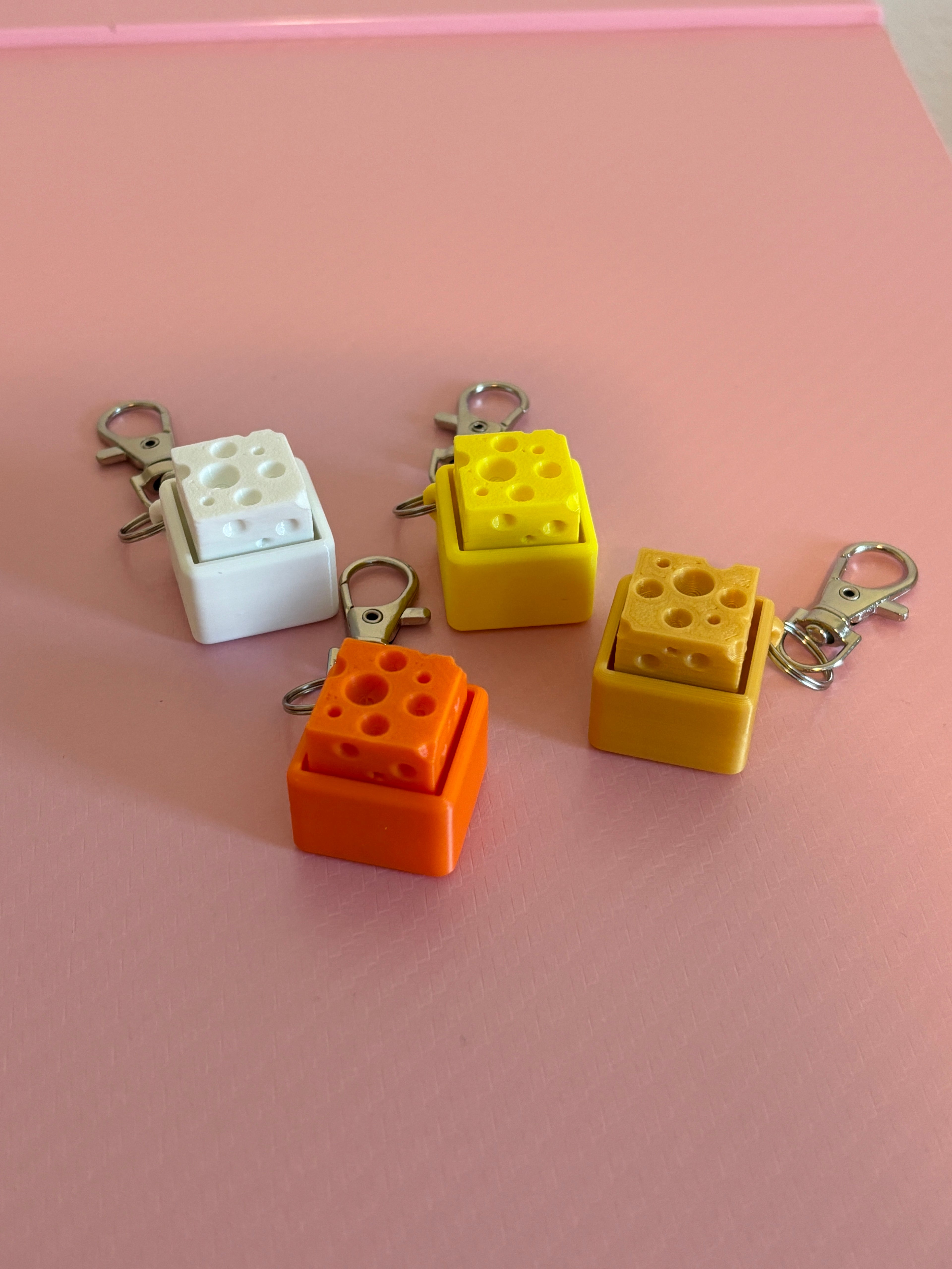 Four miniature cheese-shaped keychain clickers in yellow, orange, and white, each attached to a silver metal lobster clasp, displayed on a pink background.