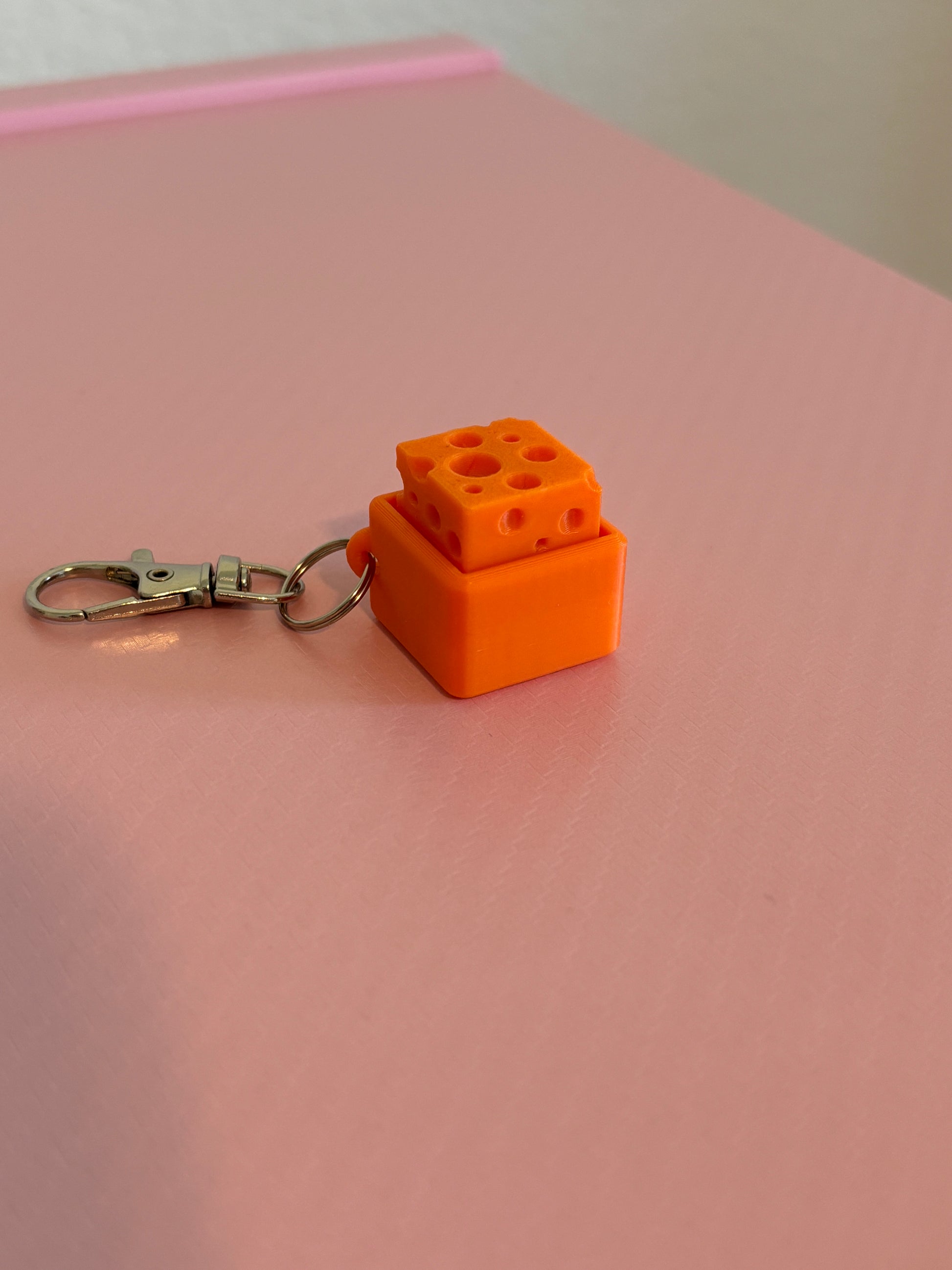 Miniature cheese-shaped keychain clicker in orange attached to a silver metal lobster clasp, displayed on a pink background.