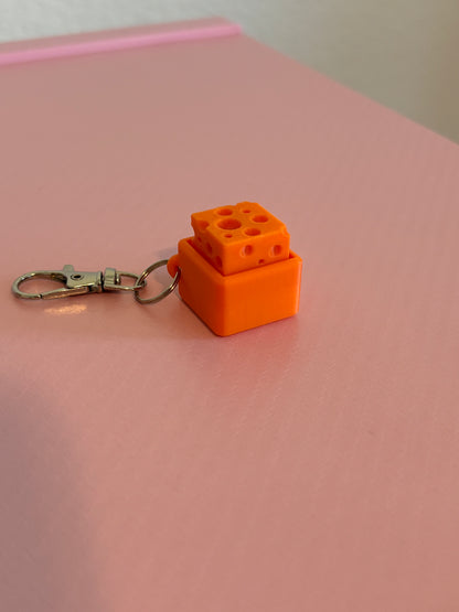 Miniature cheese-shaped keychain clicker in orange attached to a silver metal lobster clasp, displayed on a pink background.