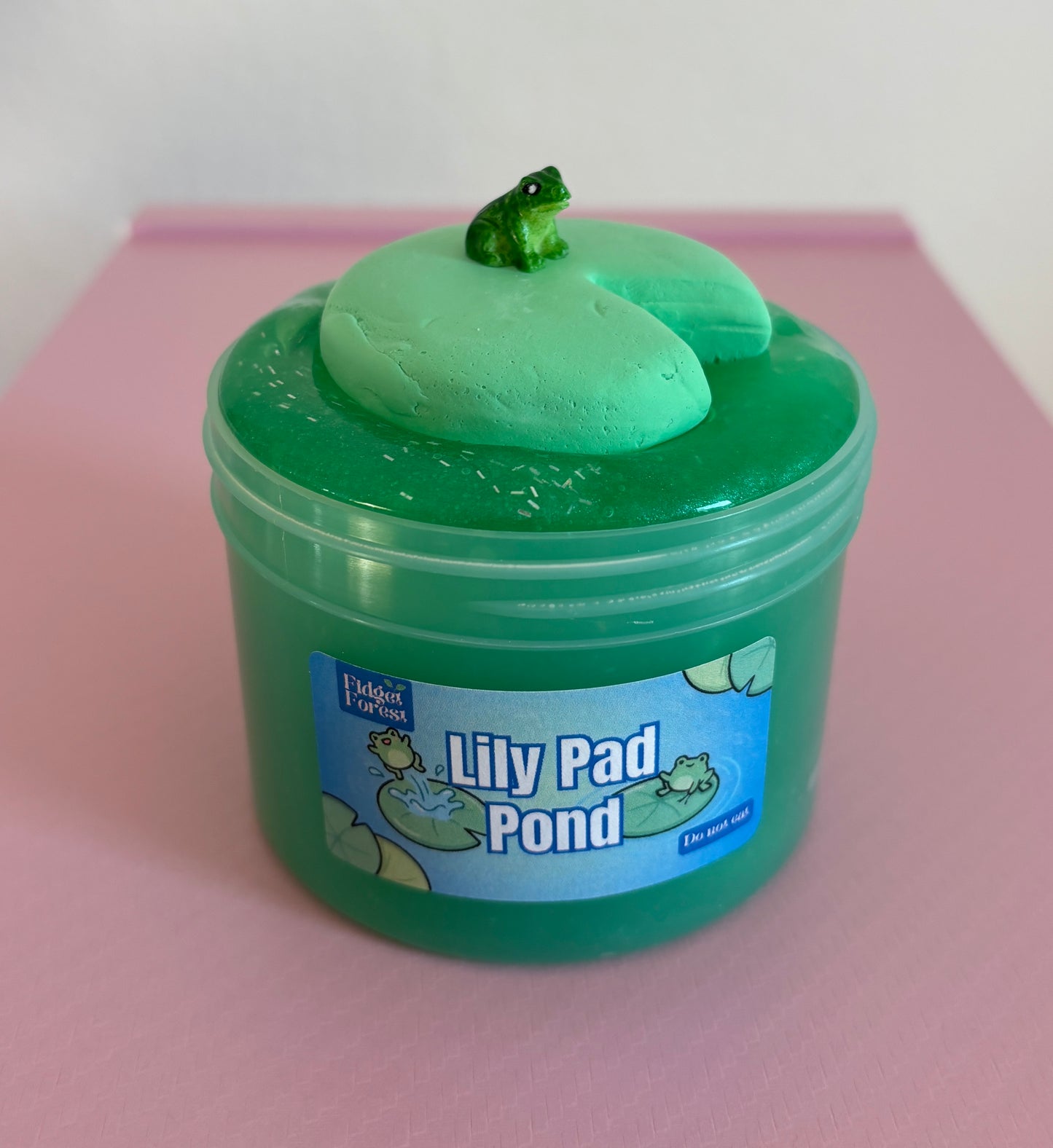 Green Jelly slime in container labeled “Lily Pad Pond” with clay lily pad and frog charm on top, in front of pink background