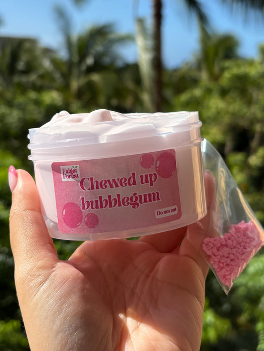 Jar of light pink slime with label that says “Chewed up bubblegum” and a bag of pink clay crumbs held by hand in front of a tropical background.