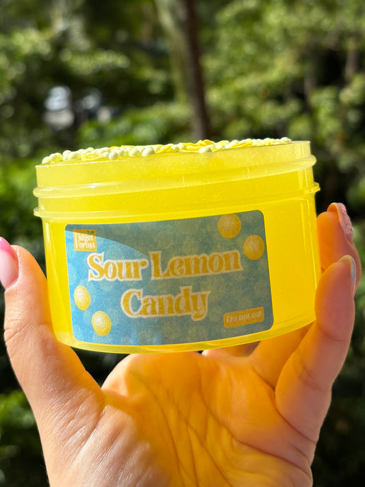 Plastic Jar of yellow slime labeled “Sour Lemon Candy” with a layer of foam beads and lemon fimo slices on top, held by a hand in front of a tropical background.