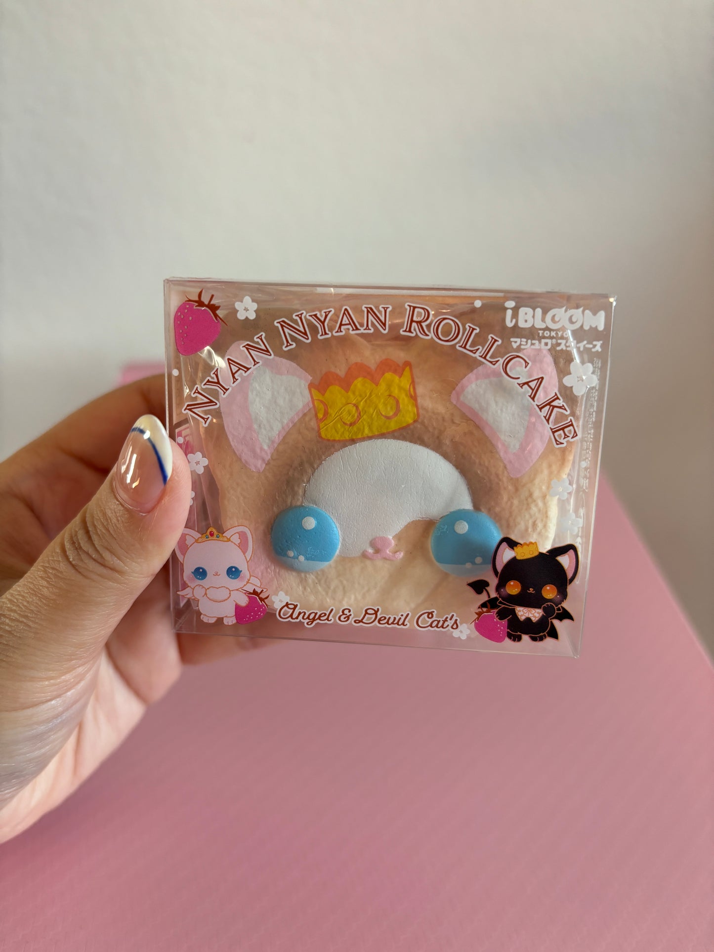 A hand holds a packaged brown cat-themed roll cake squishy toy labeled 'Nyan Nyan Roll Cake' with 'Angel & Devil Cats' and 'i BLOOM' branding, against a pink background.