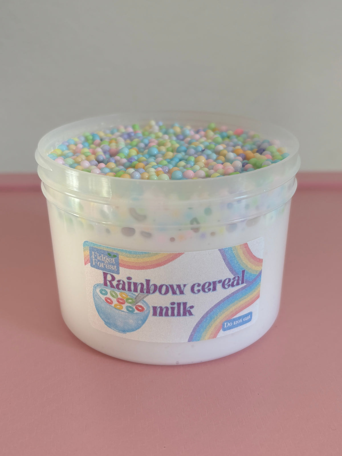 Container of white slime with pastel rainbow foam beads on top labeled ‘rainbow cereal milk’ with a pink background