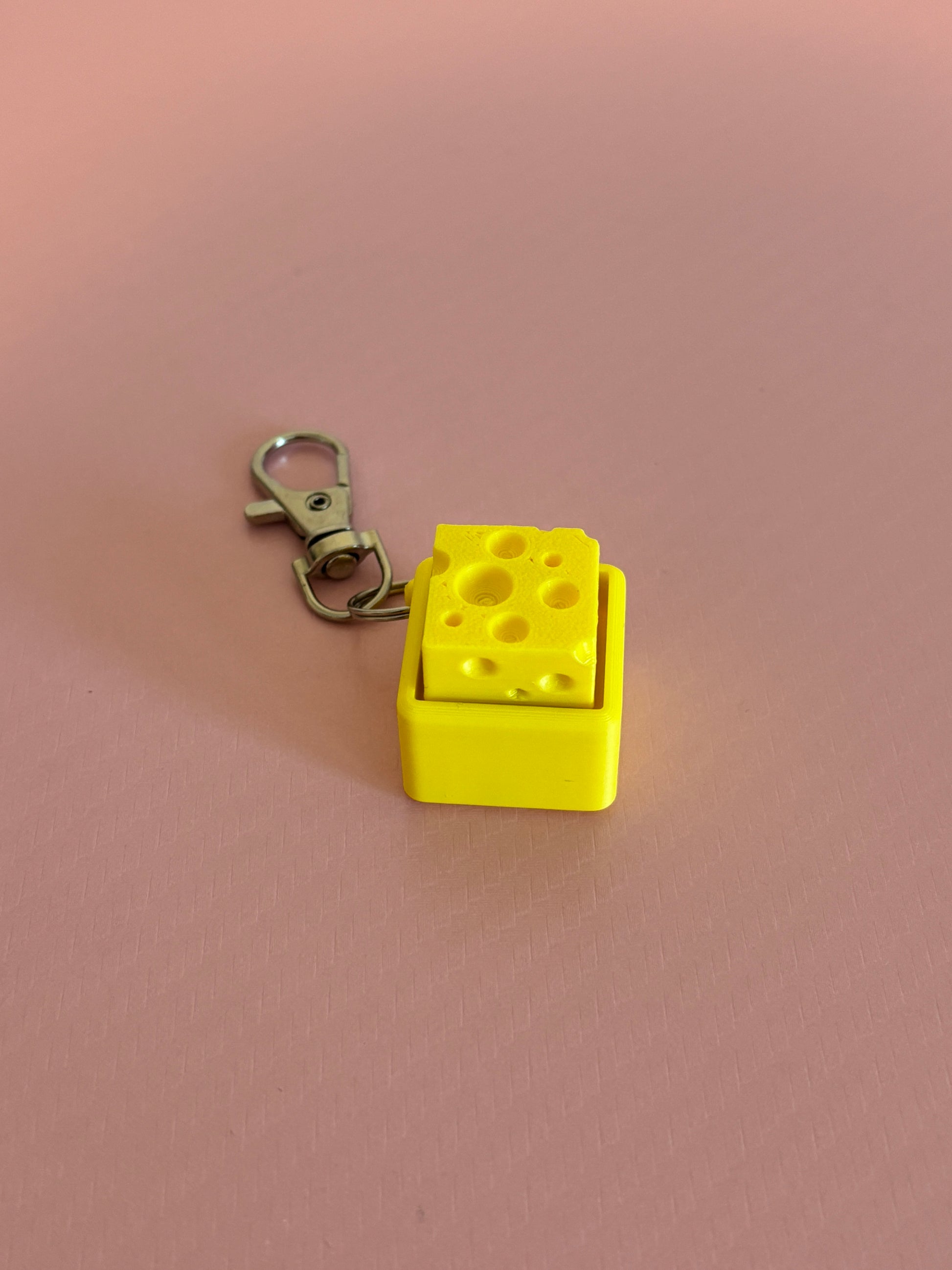Miniature cheese-shaped keychain clicker in yellow attached to a silver metal lobster clasp, displayed on a pink background.