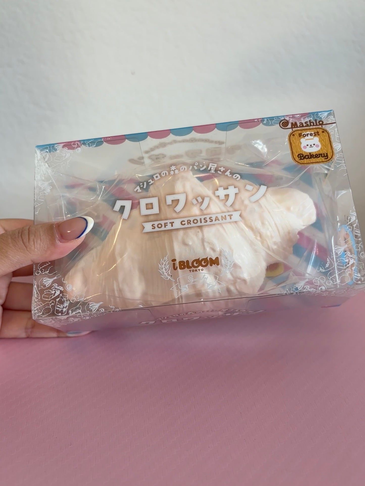 A hand holding a clear plastic package containing a white soft croissant-shaped squishy with Japanese text and a small gold seal, set against a light gray wall and pink surface.