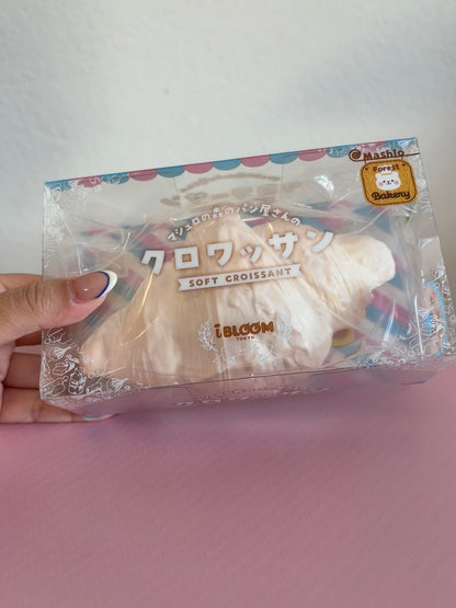 A hand holding a clear plastic package containing a white soft croissant-shaped squishy with Japanese text and a small gold seal, set against a light gray wall and pink surface.