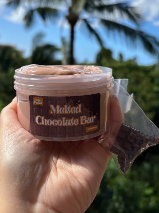 A hand holding a clear plastic jar of brown slime with a label that says “melted chocolate bar”, alongside a small clear bag of brown clay crumbs, set against a blurred tropical background with palm trees and blue sky.