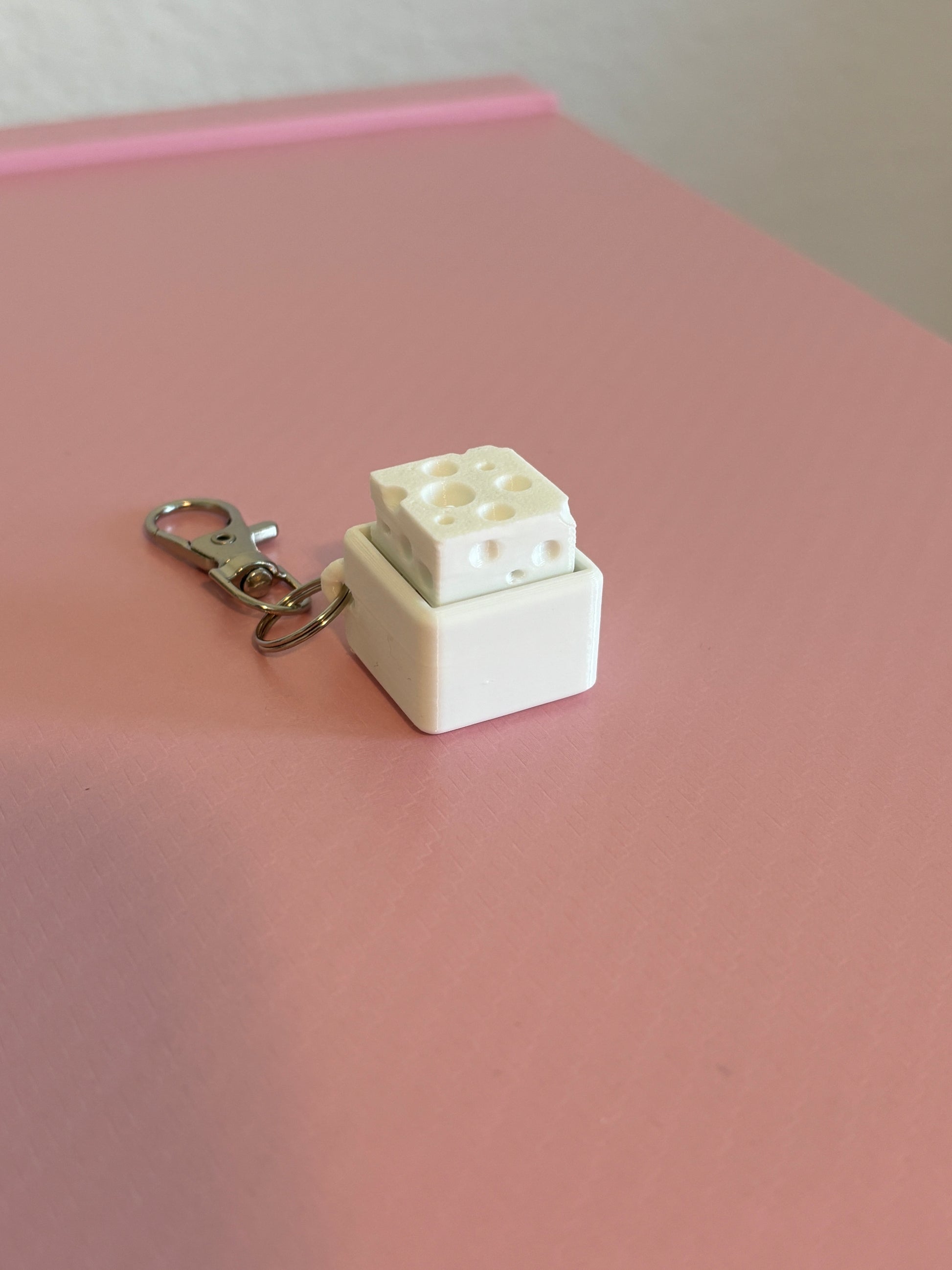 Miniature cheese-shaped keychain clicker in white attached to a silver metal lobster clasp, displayed on a pink background.