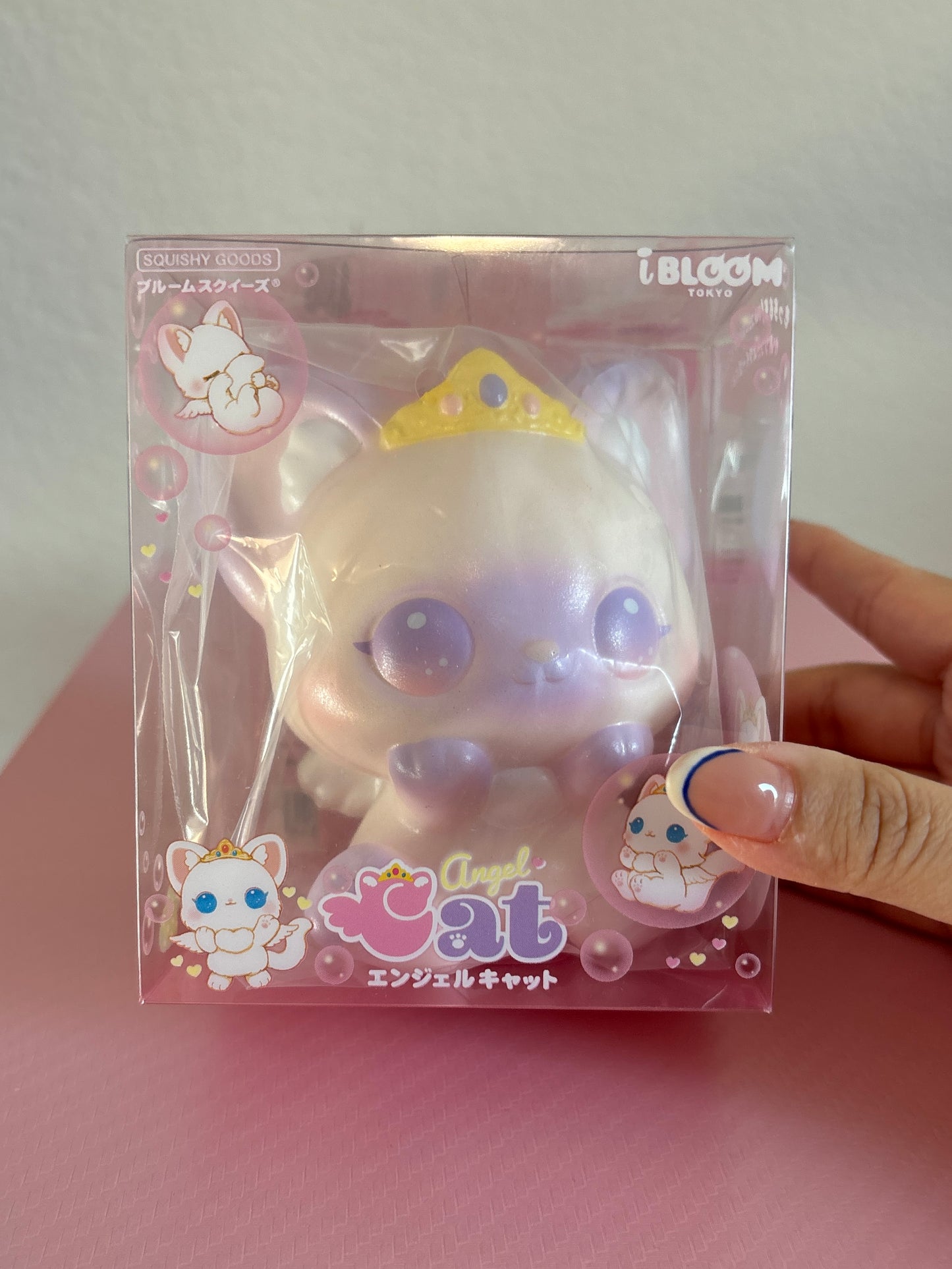 A packaged squishy toy cat with a white body, iridescent lavender-purple shading, and a small gold crown, shown inside a clear plastic box with the words “Angel Cat”, and Japanese text. A hand is holding the plastic box.