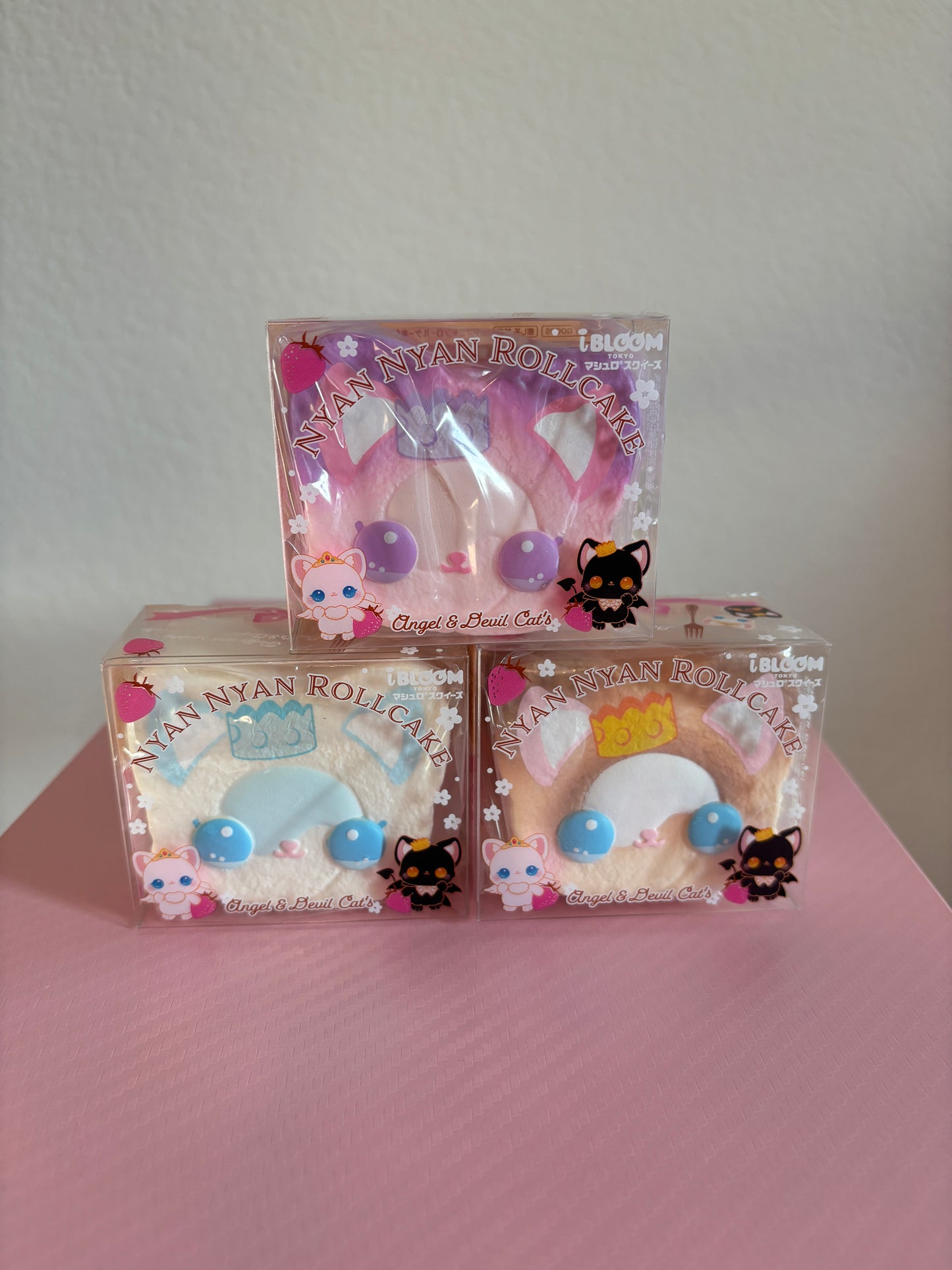 Three packaged Nyan Nyan Roll-Cat squishies stacked on a pink surface against a light wall. The top package contains a pink squishy with purple circular accents, and the two bottom packages contain brown and white-colored squishies with blue and yellow accents.