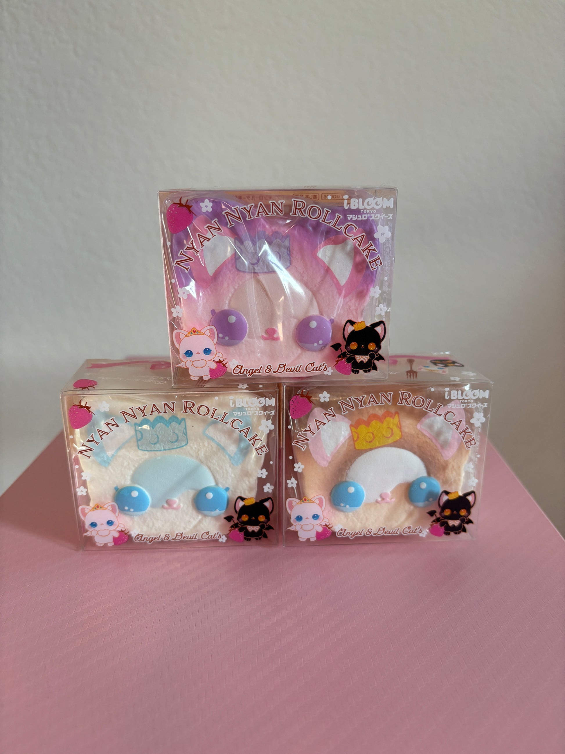 Three packaged Nyan Nyan Roll-Cat squishies stacked on a pink surface against a light wall. The top package contains a pink squishy with purple circular accents, and the two bottom packages contain brown and white-colored squishies with blue and yellow accents.