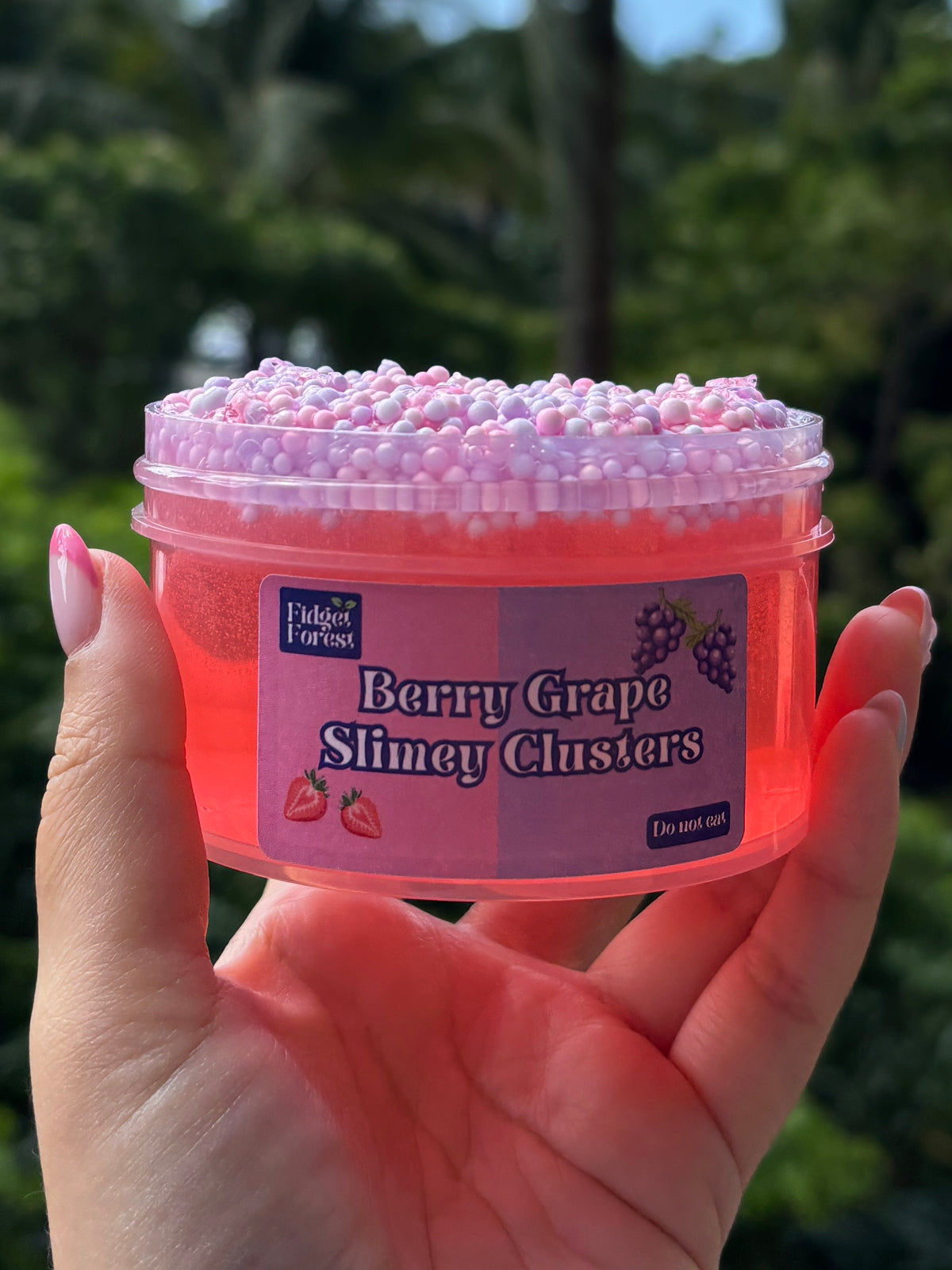 Jar of red clear slime with purple and pink foam bead layer at the top. The slime is by Fidget Forest and the label says “Berry Grape Slimey Clusters” and “Do not eat” and is being held by hand in front of tropical background.