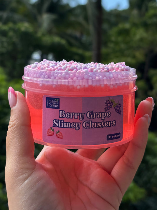 Jar of red clear slime with purple and pink foam bead layer at the top. The slime is by Fidget Forest and the label says “Berry Grape Slimey Clusters” and “Do not eat” and is being held by hand in front of tropical background.