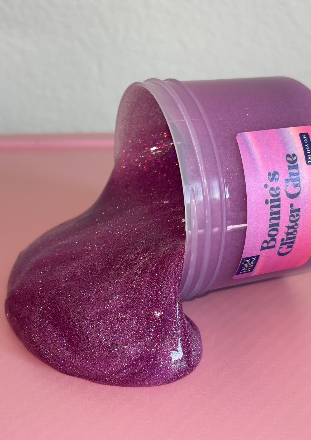 Purple Holographic Glitter slime spilling out of container container labeled “Bonnie’s Glitter Glue” with pink background 