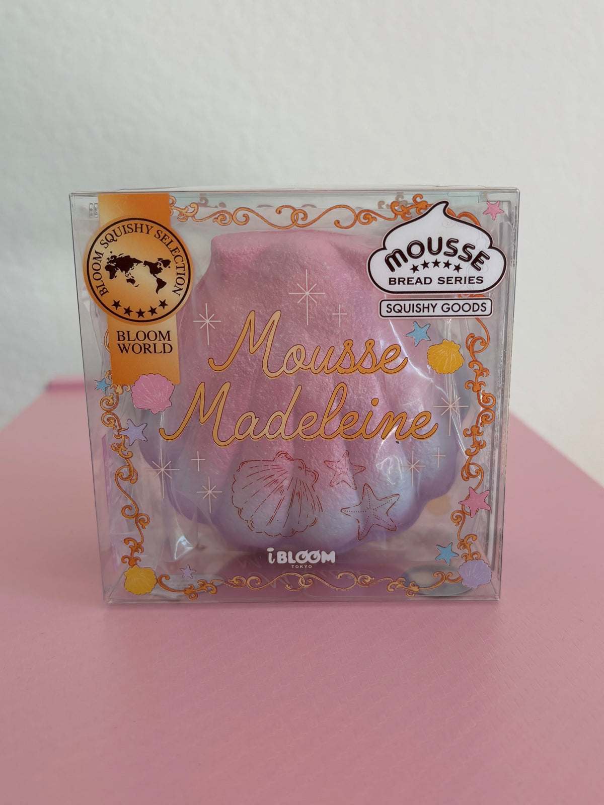 A hand holding a clear plastic package containing a shimmery pink, purple, and blue squishy toy shaped like a madeleine cookie, with decorative gold foil text and branding including 'Mousse Madeleine' by the brand IBLOOM.