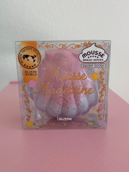 A hand holding a clear plastic package containing a shimmery pink, purple, and blue squishy toy shaped like a madeleine cookie, with decorative gold foil text and branding including 'Mousse Madeleine' by the brand IBLOOM.