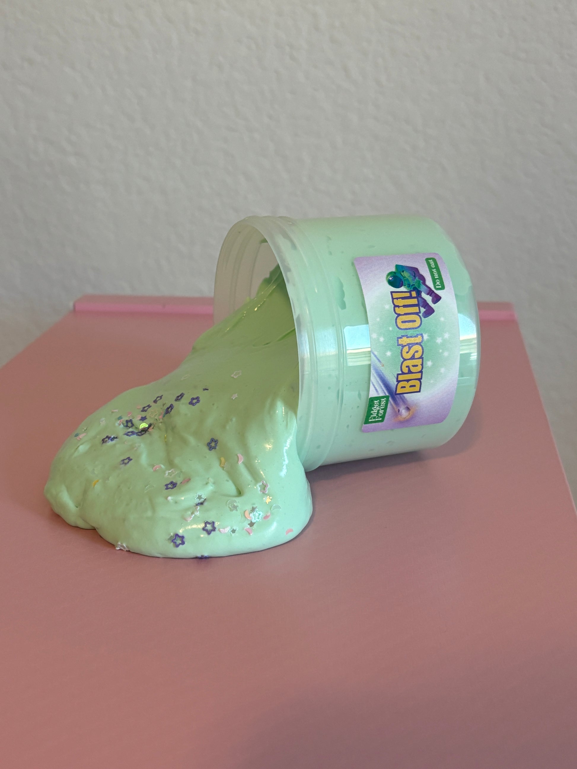 Green and White Slime spilling out of container with variety of purple and white star glitter, and moon glitter, in front of pink background