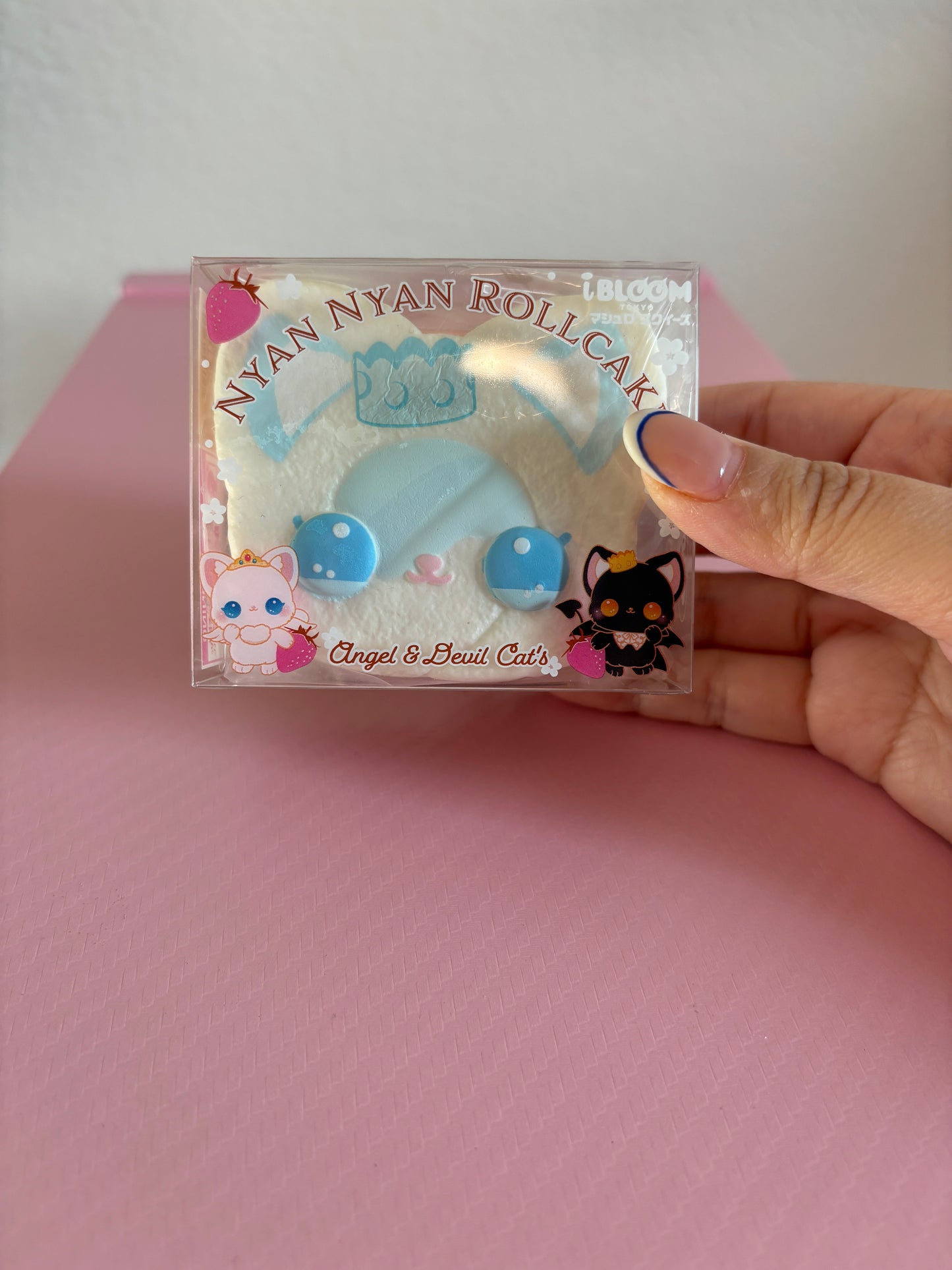 A hand holds a packaged white and blue cat-themed roll cake squishy toy labeled 'Nyan Nyan Roll Cake' with 'Angel & Devil Cats' and 'i BLOOM' branding, against a pink background.