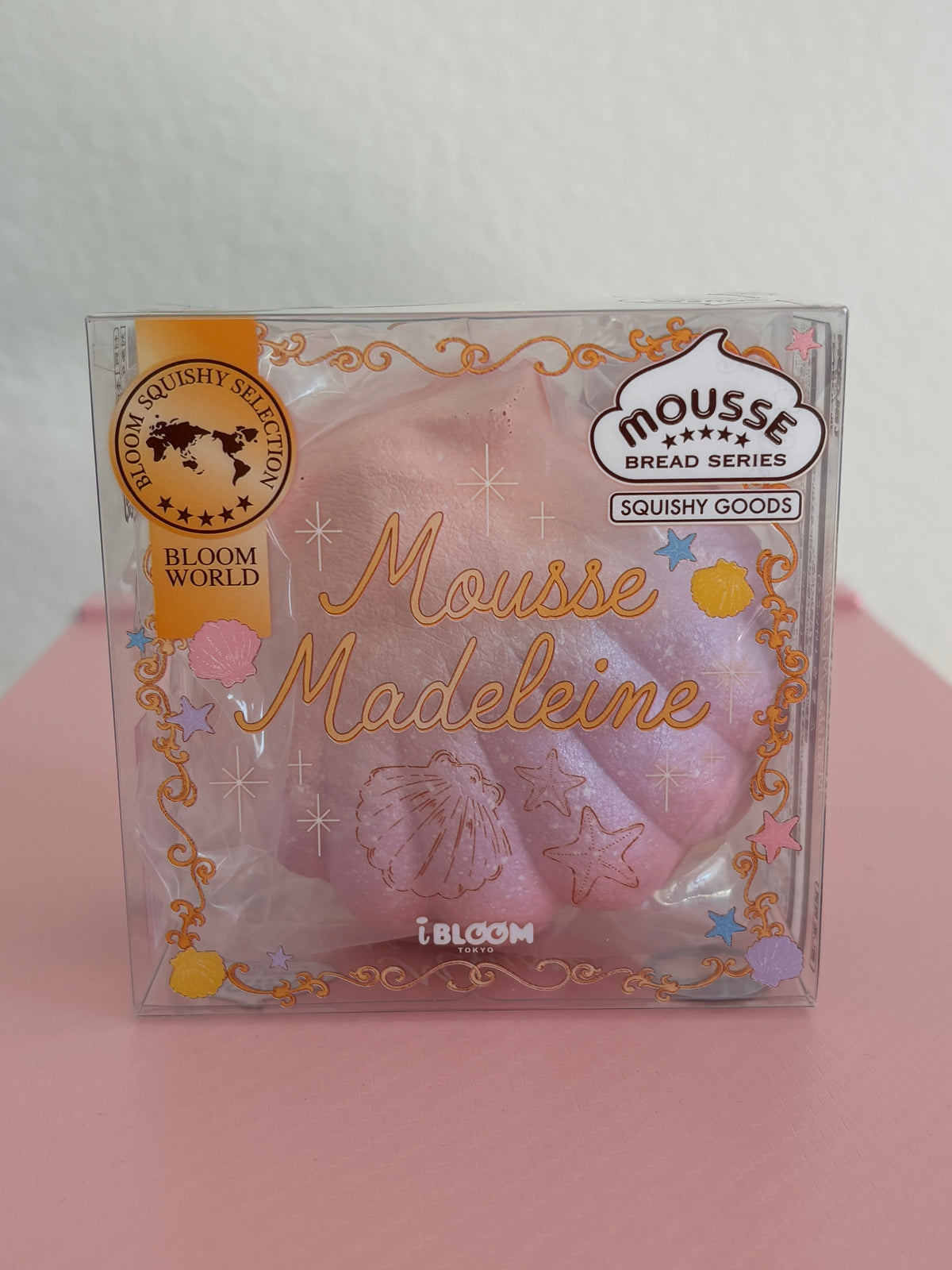 A hand holding a clear plastic package containing a shimmery purple and pink squishy toy shaped like a madeleine cookie, with decorative gold foil text and branding including 'Mousse Madeleine' by the brand IBLOOM.