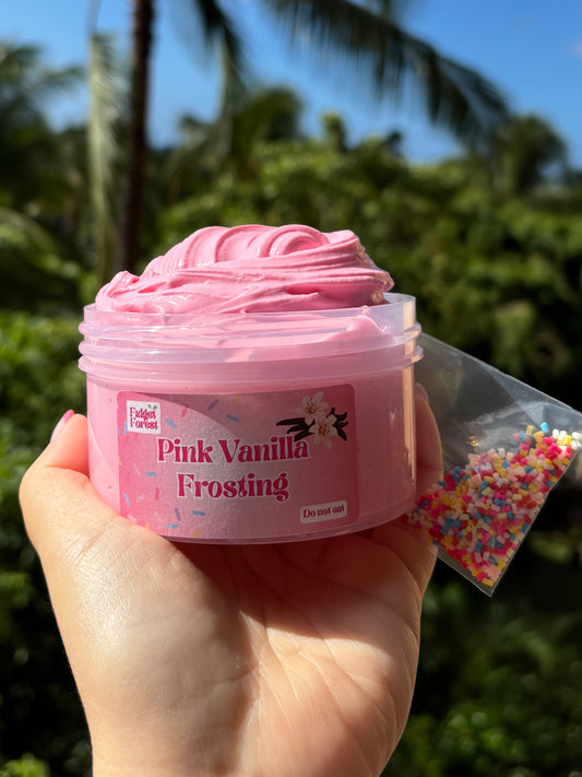 Jar of pink slime with label that says “Pink Vanilla Frosting” and a bag of rainbow sprinkles held by a hand in front of a tropical background.