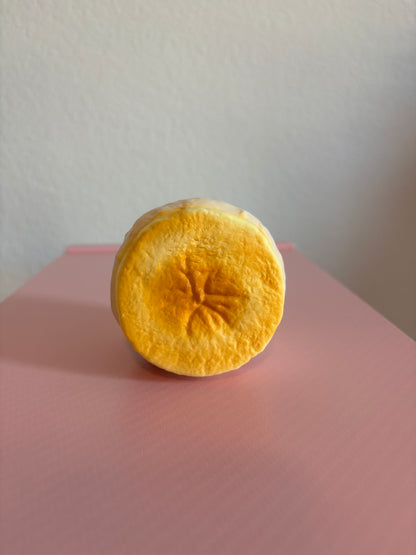 Hachimi Banana Cream Puff Squishy toy showing the round slice face with a vibrant golden-yellow caramelized finish, a star shaped center crease, and a pale cream-colored outer pastry shell.  Centered on a white and pink background.