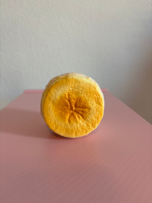 Hachimi Banana Cream Puff Squishy toy showing the round slice face with a vibrant golden-yellow caramelized finish, a star shaped center crease, and a pale cream-colored outer pastry shell.  Centered on a white and pink background.