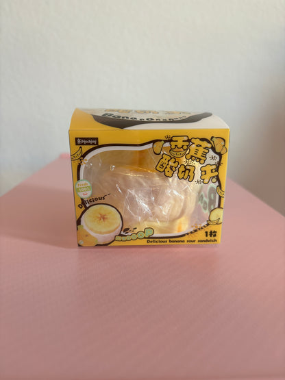 Hachimi Banana Cream Puff Squishy toy inside its yellow retail display box, featuring banana graphics, stylized Chinese characters, and a clear font window showing the packaged toy. Centered on a white and pink background.