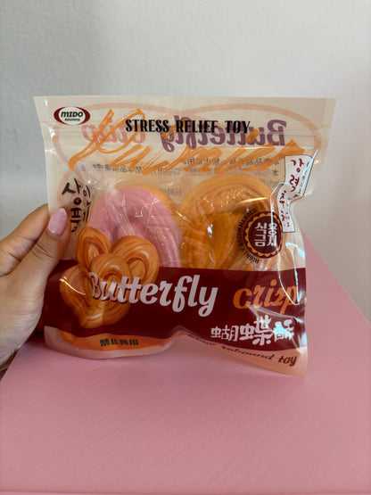 Mido Butterfly Crisp Squishy in its clear retail packaging, showing one slow-rising foam pastry shaped like a realistic butterfly palmier cookie with one half covered in pastel pink frosting. Held by a hand on a white and pink background.