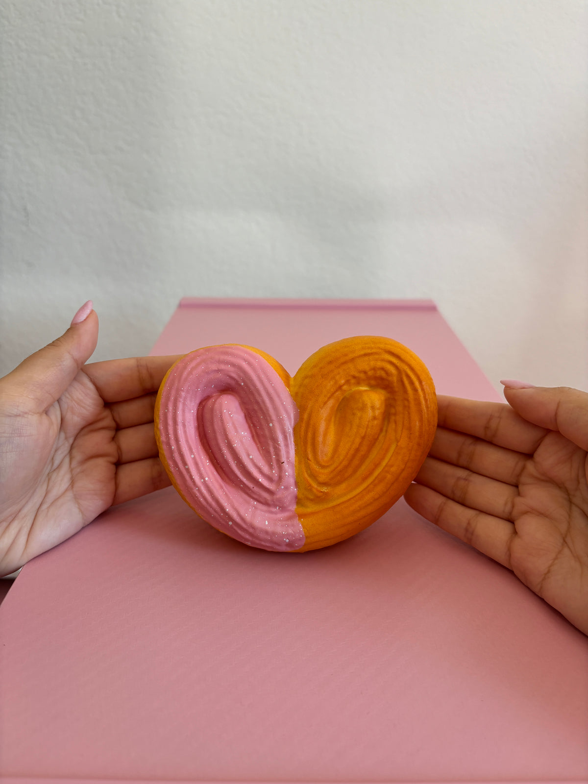 2 hands holding heart shaped squishy by fingertips with palms facing the camera centered on a pink and white background. Squishy is shaped as a heart with a palmier visual texture on the top. The back is a smooth texture. The left vertical half of the heart has pink frosting with white speckles. The other half does not have frosting. The color of the squishy (not including the pink frosting) is a burnt orange color.