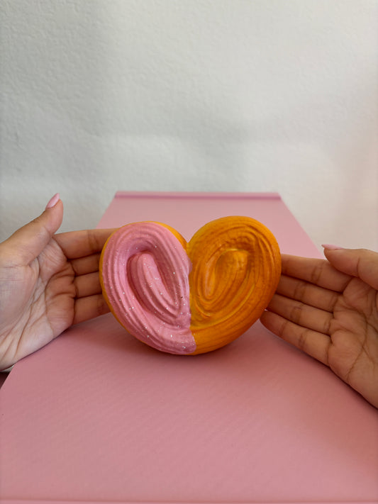 2 hands holding heart shaped squishy by fingertips with palms facing the camera centered on a pink and white background. Squishy is shaped as a heart with a palmier visual texture on the top. The back is a smooth texture. The left vertical half of the heart has pink frosting with white speckles. The other half does not have frosting. The color of the squishy (not including the pink frosting) is a burnt orange color.