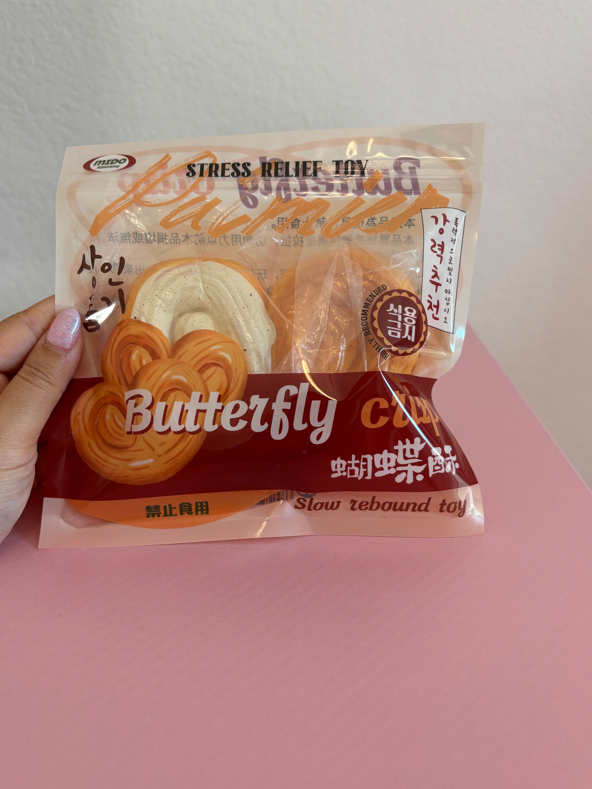 Mido Butterfly Crisp Squishy in its clear retail packaging, showing one slow-rising foam pastry shaped like a realistic butterfly palmier cookie with one half covered in white frosting. Held by a hand on a white and pink background.