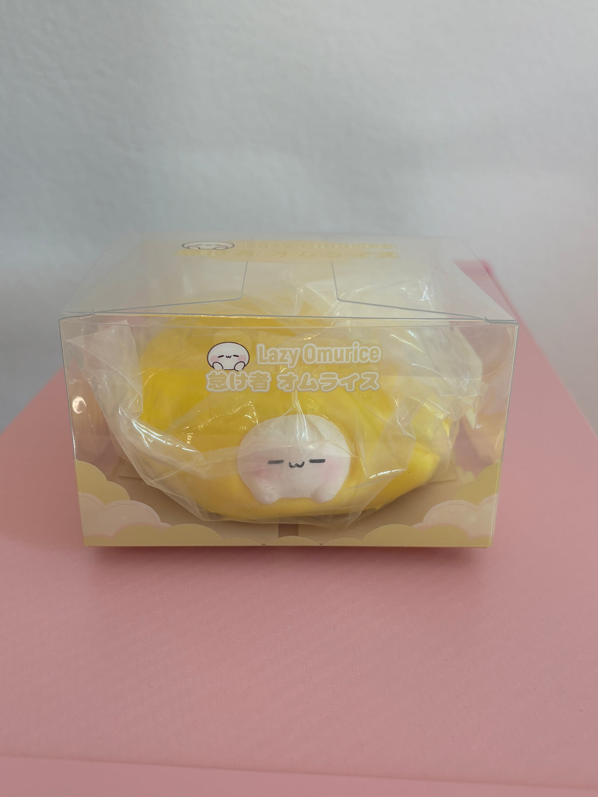 Yellow Omurice squishy toy in its clear retail display box, featuring a cute, white character with a sleepy expression tucked into a bright yellow egg omelet. Sitting centered on a pink and white background.