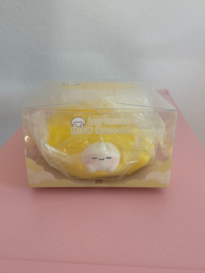 Yellow Omurice squishy toy in its clear retail display box, featuring a cute, white character with a sleepy expression tucked into a bright yellow egg omelet. Sitting centered on a pink and white background.