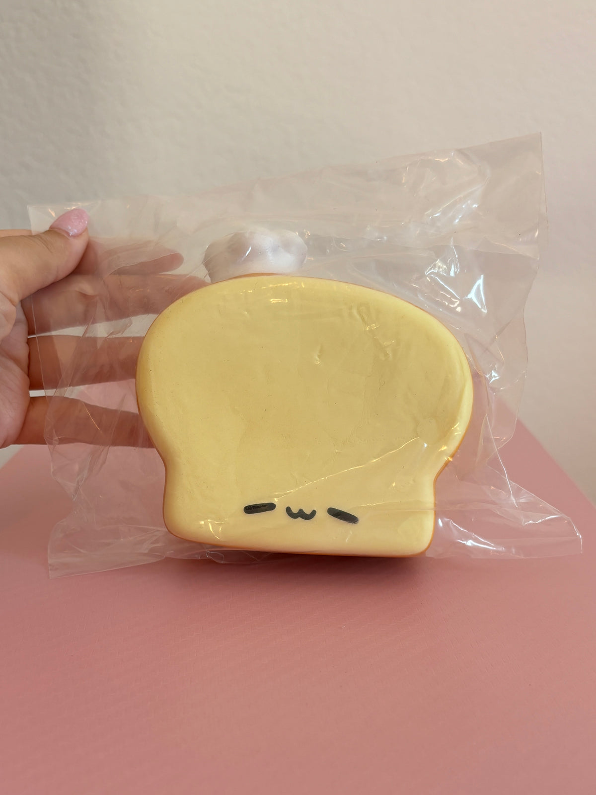 Nunu Lazy Toast Squishy toy inside its clear plastic packaging, featuring a thick, cream-colored slice of break with a light brown crust, a small white chef hat on its top left, and a sleepy facial expression. Held by a hand on a white and pink background.