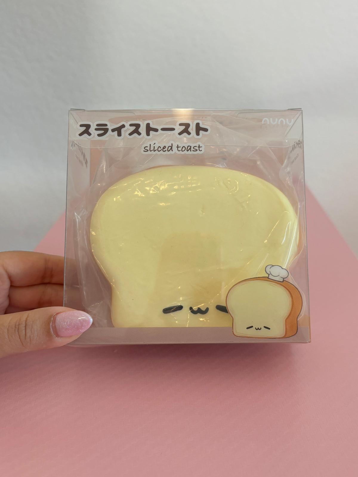 Nunu Lazy Toast Squishy toy inside its clear plastic retail display box, featuring a thick, cream-colored slice of break with a light brown crust, a small white chef hat on its top left, and a sleepy facial expression. Centered on a white and pink background. The display box has Japanese writing on the top left.