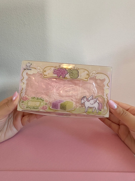A person holds a San Pink Jumbo Bread Squishy in its retail box packaging with cute unicorn and pastel illustrations. The box is clear plastic, showing the squishy inside. 
