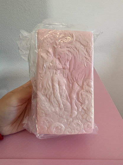 San Pink Jumbo Pony Bread Squishy toy inside clear plastic packaging, featuring a thick rectangular block embossed with an intricate carving of a mythical woman kissing the nose of a horse. Both are wrapped in swirling clouds. Held by a hand on a white and pink background.